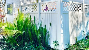 Una casa bianca con un recinto di legno bianco, circondata da piante verdi rigogliose e una decorazione a forma di farfalla.