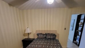 Un dormitorio con cama, mesita de noche, lámpara, ventilador de techo y alfombra estampada.