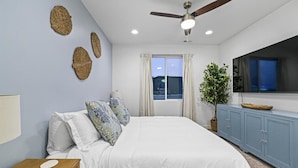 A bedroom with a bed, a ceiling fan, a window with curtains, a plant, and a TV on a cabinet.