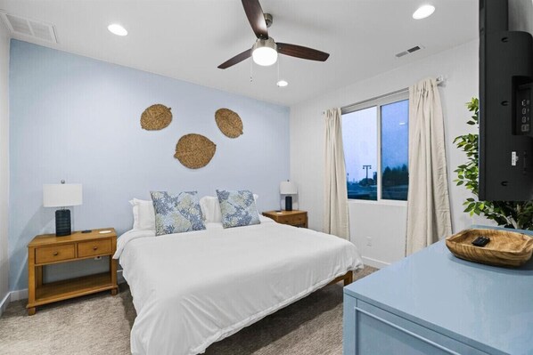 Une chambre à coucher comprenant un lit, deux tables de chevet, un ventilateur de plafond, une fenêtre avec des rideaux et un téléviseur posé sur un meuble.