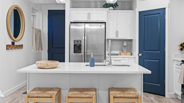 A modern kitchen with white cabinets, a stainless steel refrigerator, a double sink, and wooden stools.