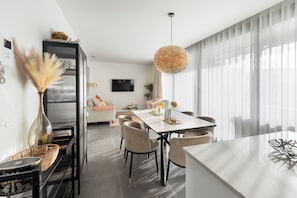 A modern living room with a dining area, featuring a sofa, a TV, and a large window with sheer curtains. - Premium Apartment with Terrace in Higuerón (Fuengirola)