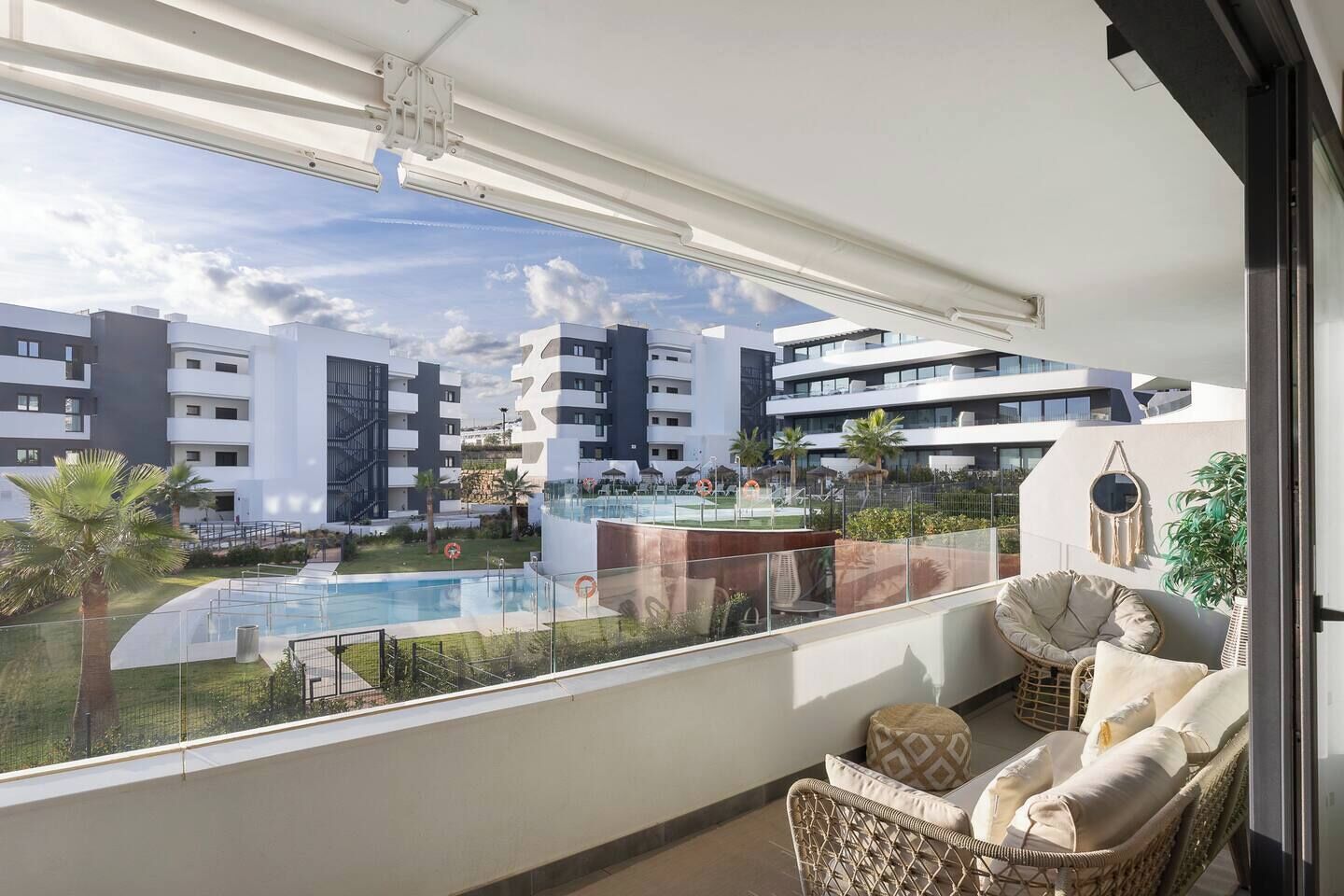A balcony with a view of a swimming pool, adjacent buildings, and a seating area with cushions.