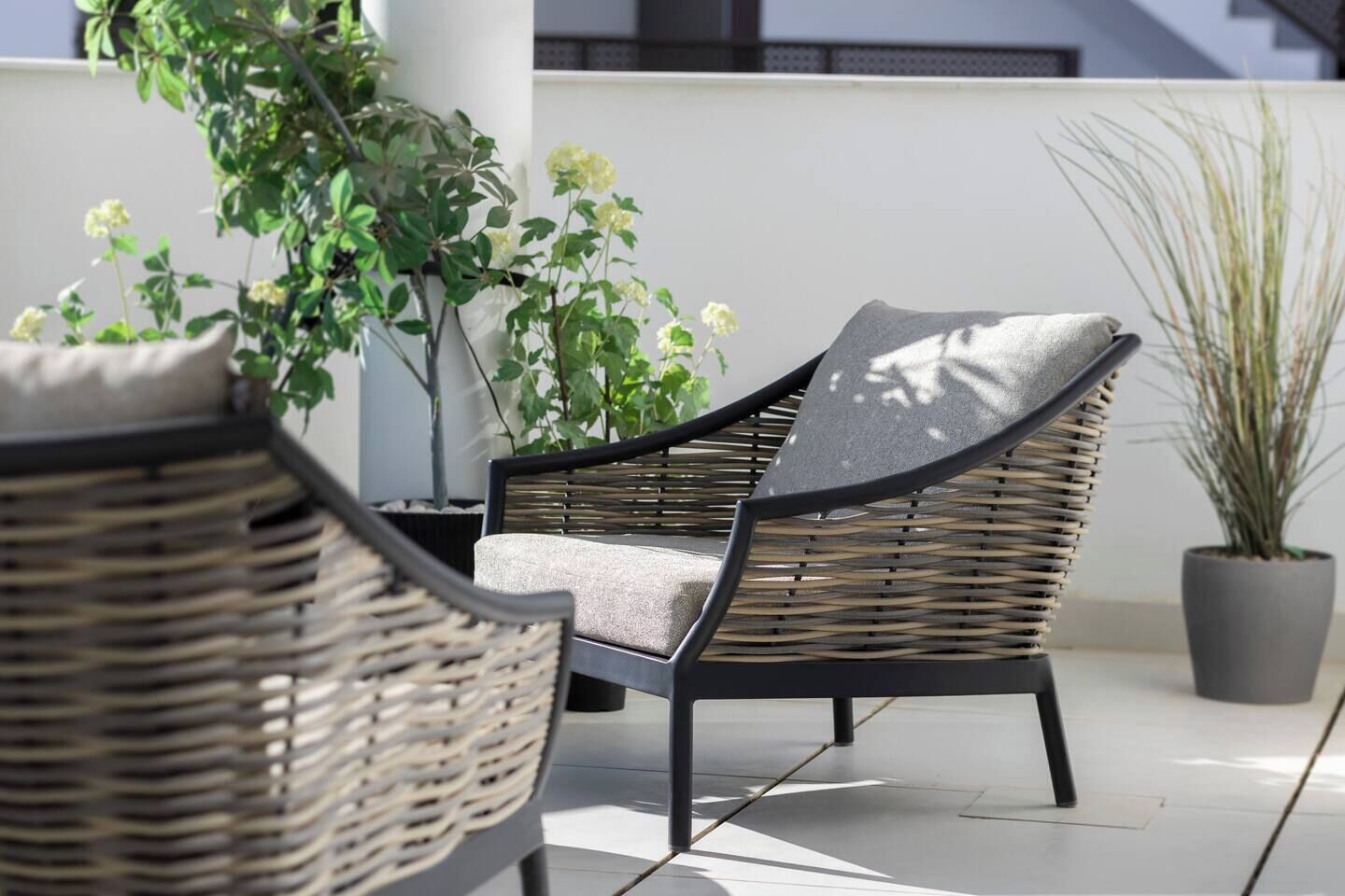 Une chaise en osier avec un coussin, placée sur une terrasse carrelée agrémentée de plantes en pot.