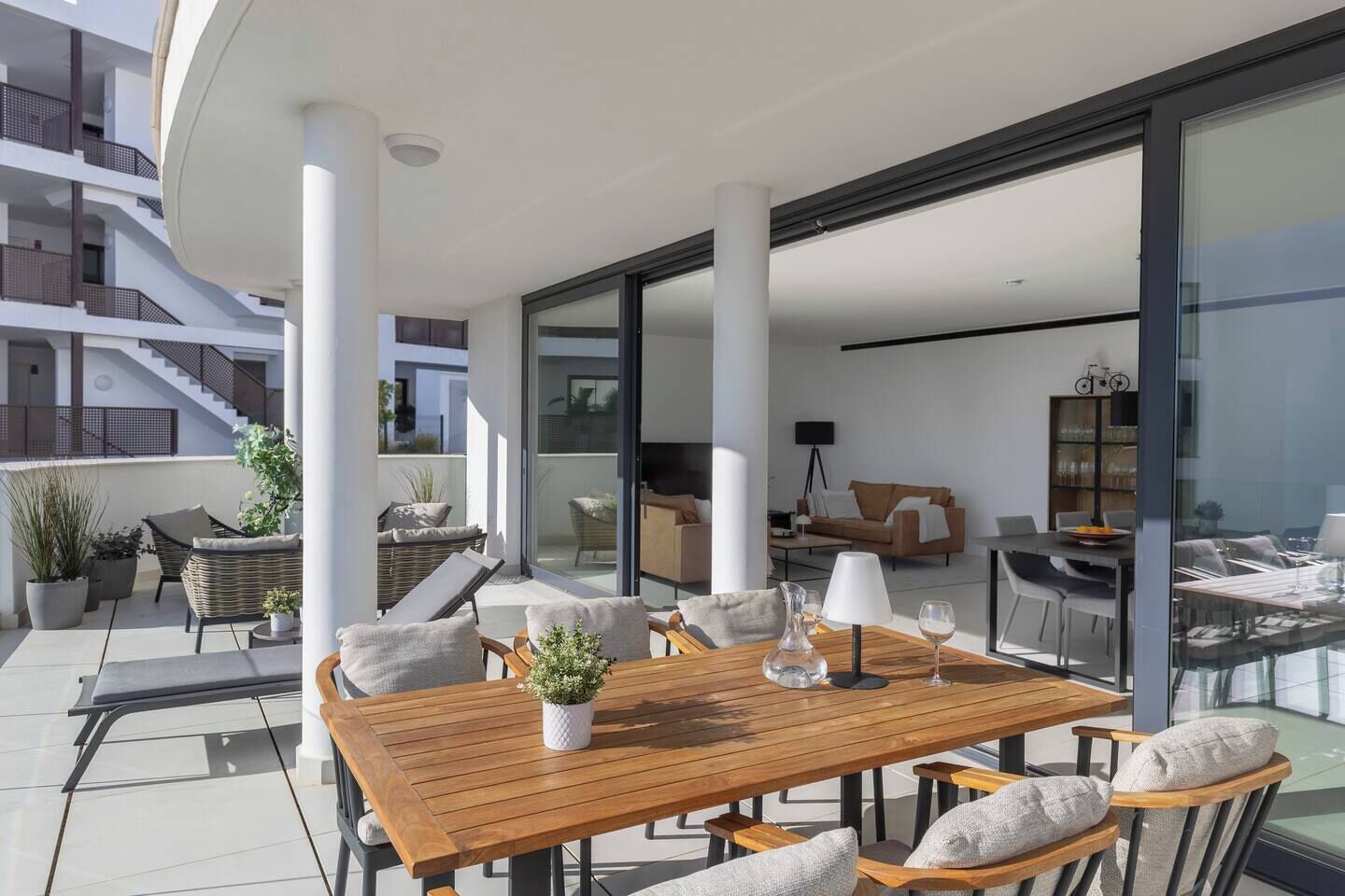 Une terrasse extérieure moderne, avec une table et des chaises en bois, entourée de plantes en pot et d’un espace de vie clos par des parois de verre.