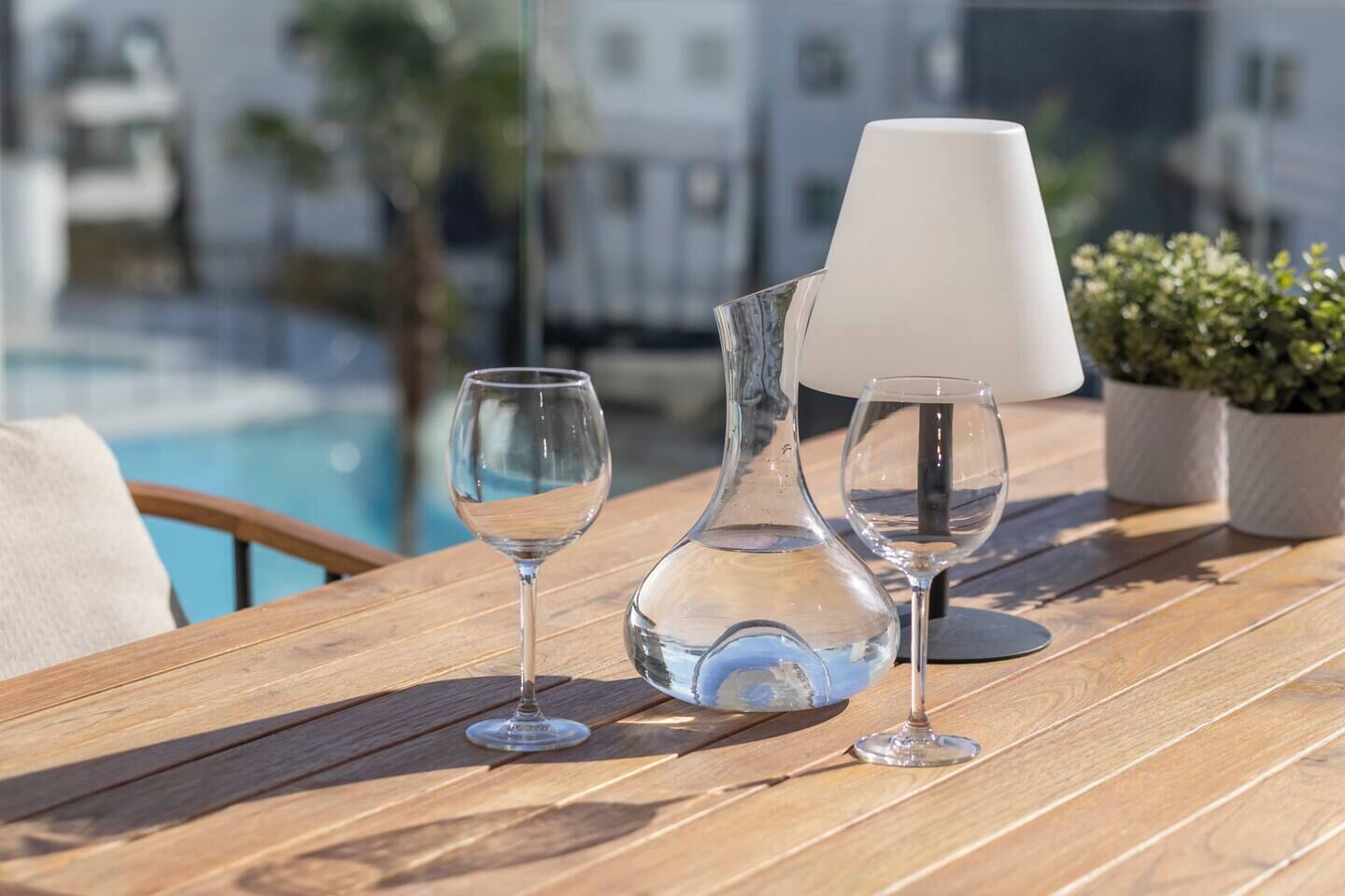 Uma mesa de madeira com duas taças de vinho e um vaso transparente, com uma piscina e edifícios ao fundo.