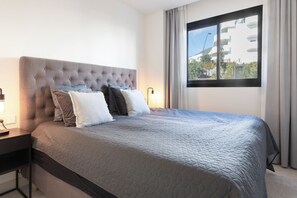 A neatly made bed with a quilted cover, a tufted headboard, and several pillows. There is a bedside table with a lamp, and a window with curtains allowing natural light. - Luxury 3-Bedroom Apartment in Higuerón (La Capellania)