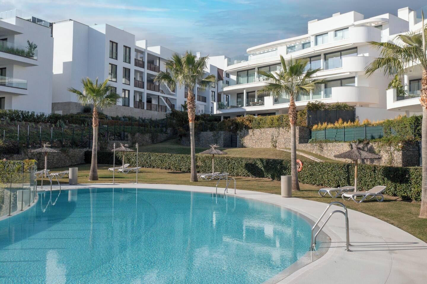 A modern pool area with clear blue water, surrounded by white buildings with balconies and palm trees.
