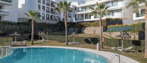 Uma área de piscina moderna com água azul cristalina, rodeada por edifícios brancos com varandas e palmeiras.