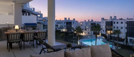 A balcony with a dining area, a sofa, and a view of a swimming pool and buildings.