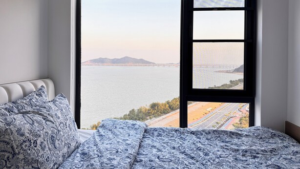 A bedroom with a large window, a bed with patterned bedding, and a view of a bridge and water.