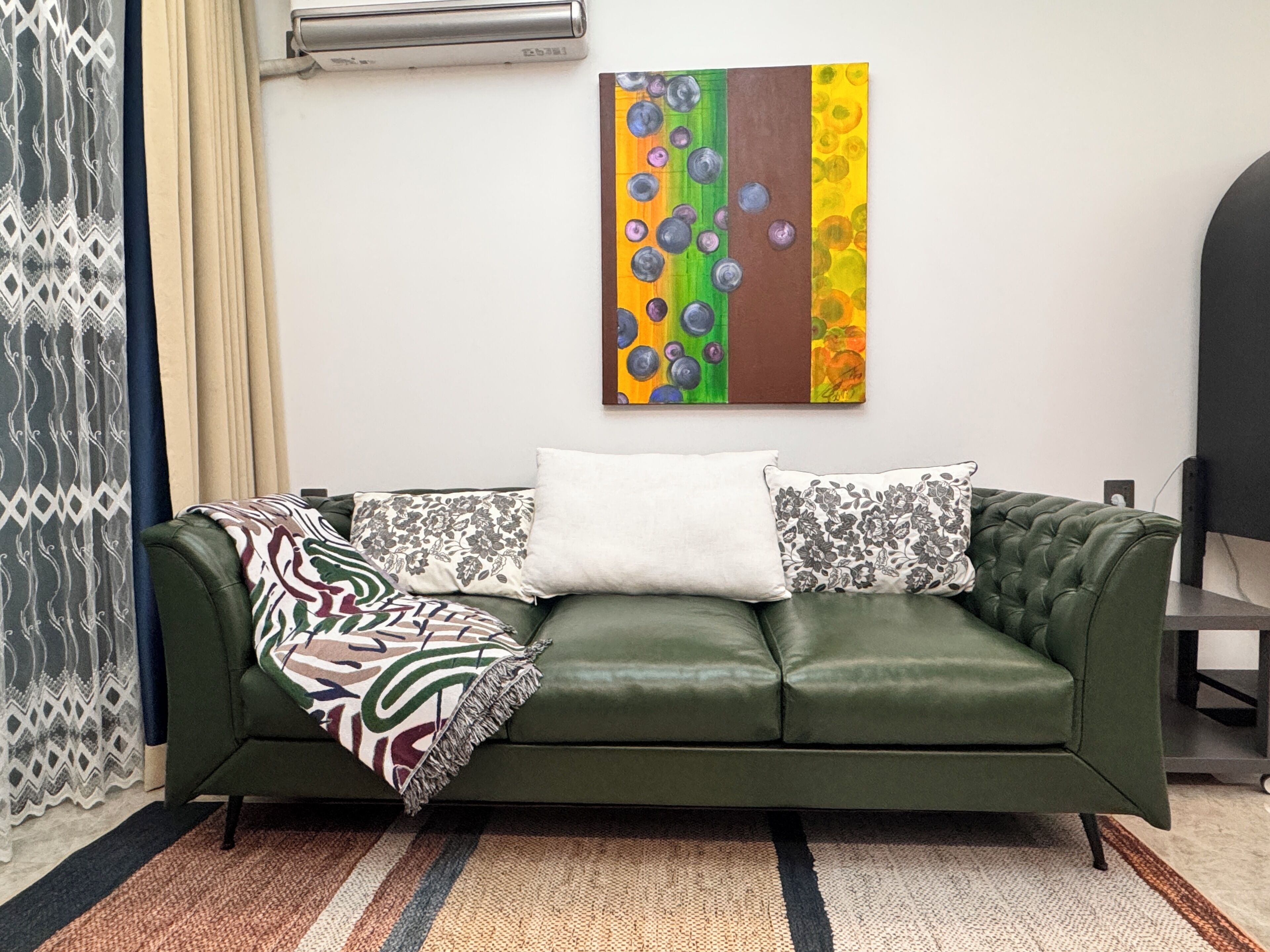 Un canapé en cuir vert agrémenté de coussins à motifs, un tableau abstrait aux couleurs vives accroché au mur et un tapis rayé sur le sol.