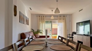 A dining area with a long wooden table, multiple chairs, woven placemats, a sofa, a television, and a potted plant.