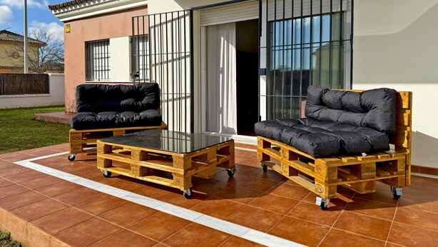 A patio with two black cushioned sofas on wooden pallets, a glass table, and a sliding glass door leading to a covered area.