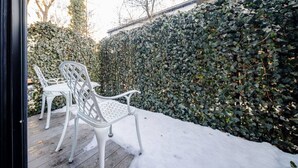 Une terrasse avec deux chaises en métal blanc et une table, entourée d’un sol recouvert de neige et d’un mur dense, entièrement couvert de lierre.