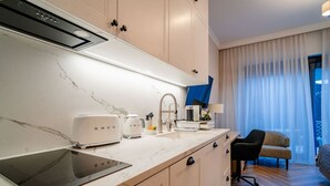 Une cuisine moderne équipée d’une cuisinière intégrée, d’un grille-pain et d’une machine à café. On y trouve un évier avec un robinet, une fenêtre avec des rideaux légers et une chaise dans un coin.