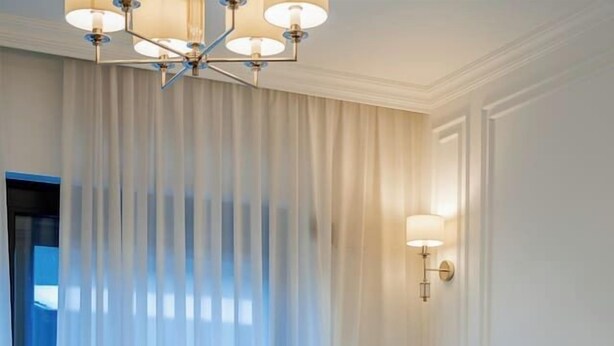 Une chambre d’hôtel avec un lit, une table de chevet sur laquelle reposent deux tasses, un lustre et une vue par une fenêtre ornée de rideaux.