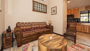 Un salon avec un canapé à motifs, une table basse ronde en bois, une petite table d’appoint et un coin cuisine doté de meubles en bois et d’un four à micro-ondes.