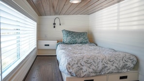 A bedroom with a bed, a nightstand, a wall-mounted lamp, and a window with blinds.