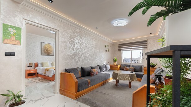 A modern living room with a sofa, a coffee table, and a dining area. There is a bed in the background and a staircase leading to an upper level. - Mina Home minutes from City Center and beach (Agadir)