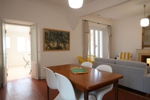 Ein Essbereich mit einem Holztisch, weißen Stühlen und einer grünen Tischmatte. Im Hintergrund befindet sich eine graue Couch und ein gelber Sessel.