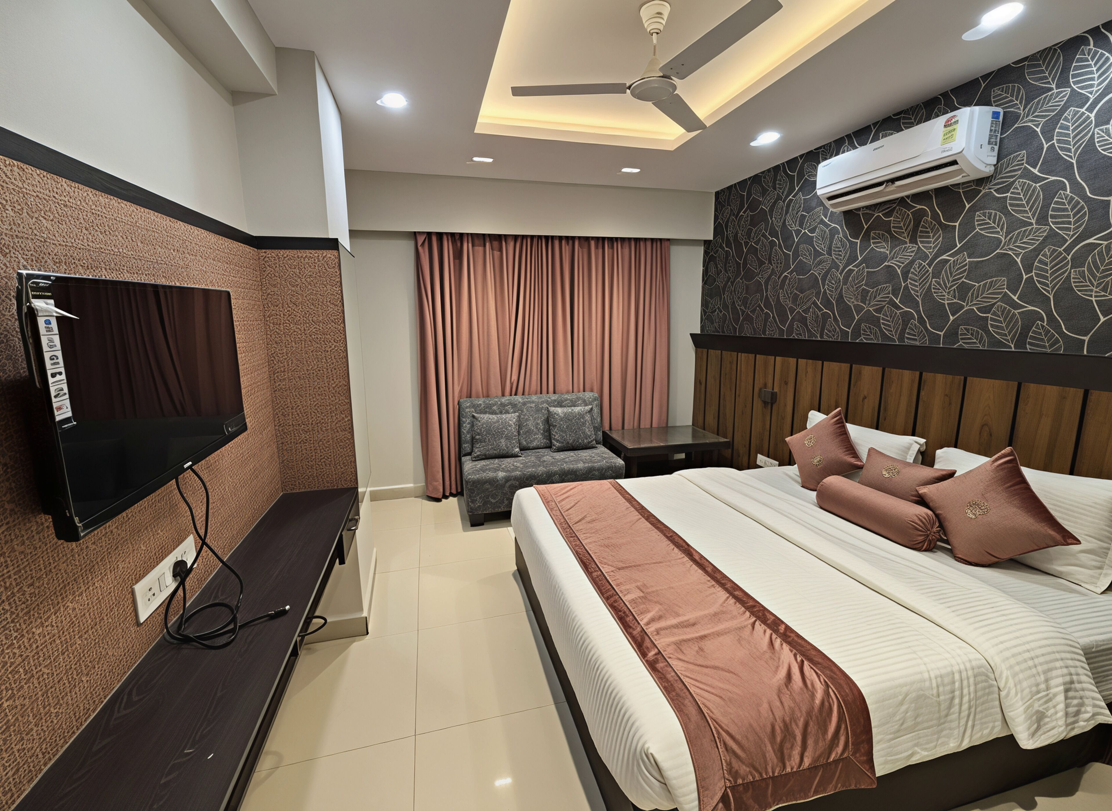 Een moderne hotelkamer met een flatscreen televisie, een bed met een bruin hoofdeinde, een grijze bank, een klein tafeltje en een airconditioning.
