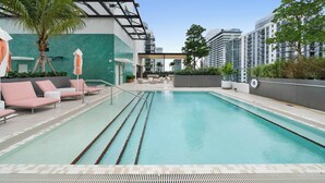 Ein Swimmingpool mit Liegestühlen und Sonnenschirmen, umgeben von Grünflächen und modernen Gebäuden.