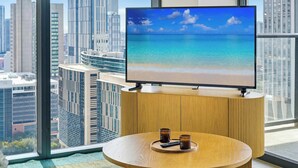 Ein modernes Zimmer mit einem großen Fernseher auf einem Holzständer, einem runden Tisch mit zwei Fernbedienungen und Blick auf eine Stadtlandschaft durch große Fenster.