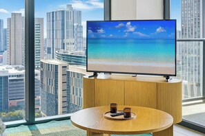 A modern room with a large TV on a wooden stand, a round table with two remote controls, and a view of a cityscape through large windows. - Downtown Drift-Luxury Amenities, and Pool (Miami)