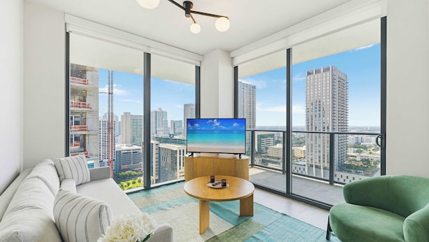 Ein modernes Wohnzimmer mit großem Fernseher, einer Couch, einem grünen Sessel und Blick auf eine Stadtlandschaft durch große Fenster.