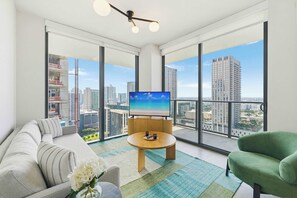 Un soggiorno moderno con una grande televisione, un divano, una poltrona verde e vista sulla città attraverso ampie finestre.