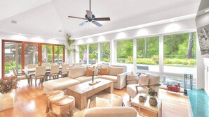 A spacious living room with a large sofa set, a coffee table, and a dining area with chairs and a table. There is a ceiling fan and large windows offering a view of the outdoors. - Waterfront Sonoma Russian Rvr Lux Vineyard Ranch on Creek, Pool+HotTub+Bocce+Pet (Sebastopol)