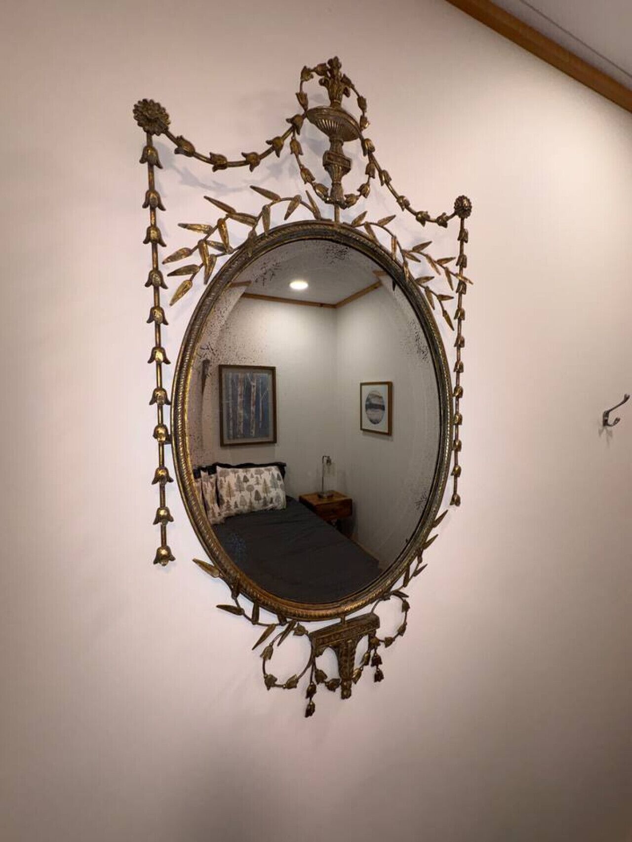 A room with a large ornate mirror on the wall, a bed with pillows, a nightstand, and two framed pictures.