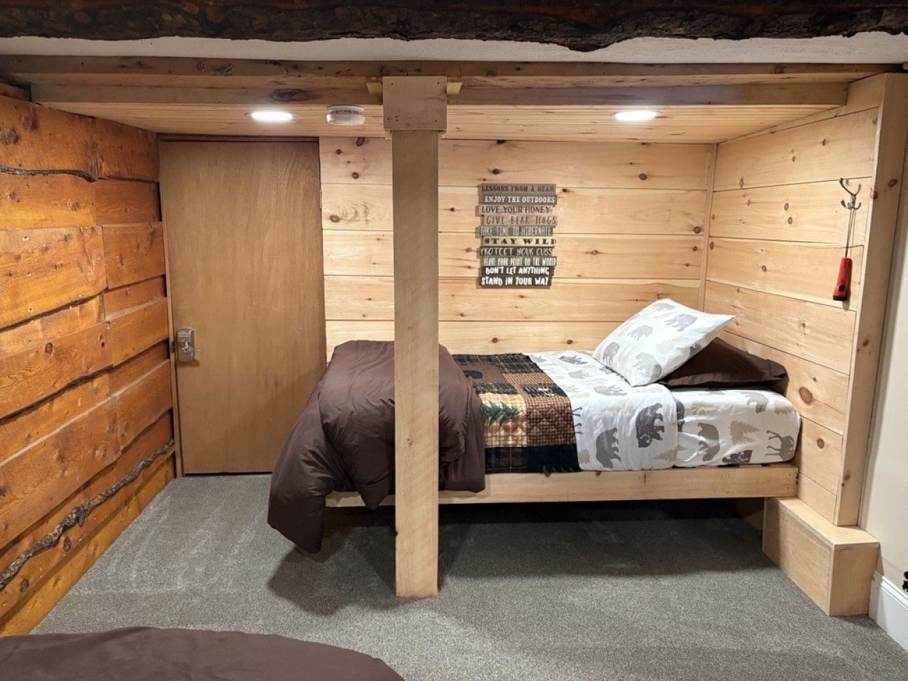 A small wooden room with a bunk bed, a door, a wall-mounted sign, and a small stool.