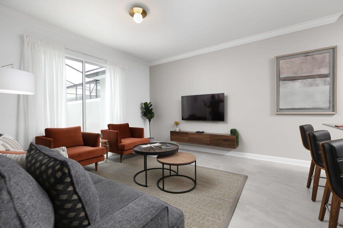 A modern living room with a grey sofa, two orange armchairs, a coffee table, and a TV on the wall.