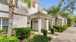 A row of modern, single-story buildings with white exteriors and brown trim, featuring a covered entrance and a well-maintained garden with various shrubs and a palm tree. - Storey Lake 4BR Townhome - 10 min to Disney (Kissimmee)