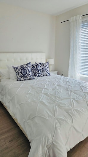 Een net opgemaakt bed met wit beddengoed en blauwe kussens met bloemenmotief, een wit hoofdbord en een nachtkastje met een lamp.