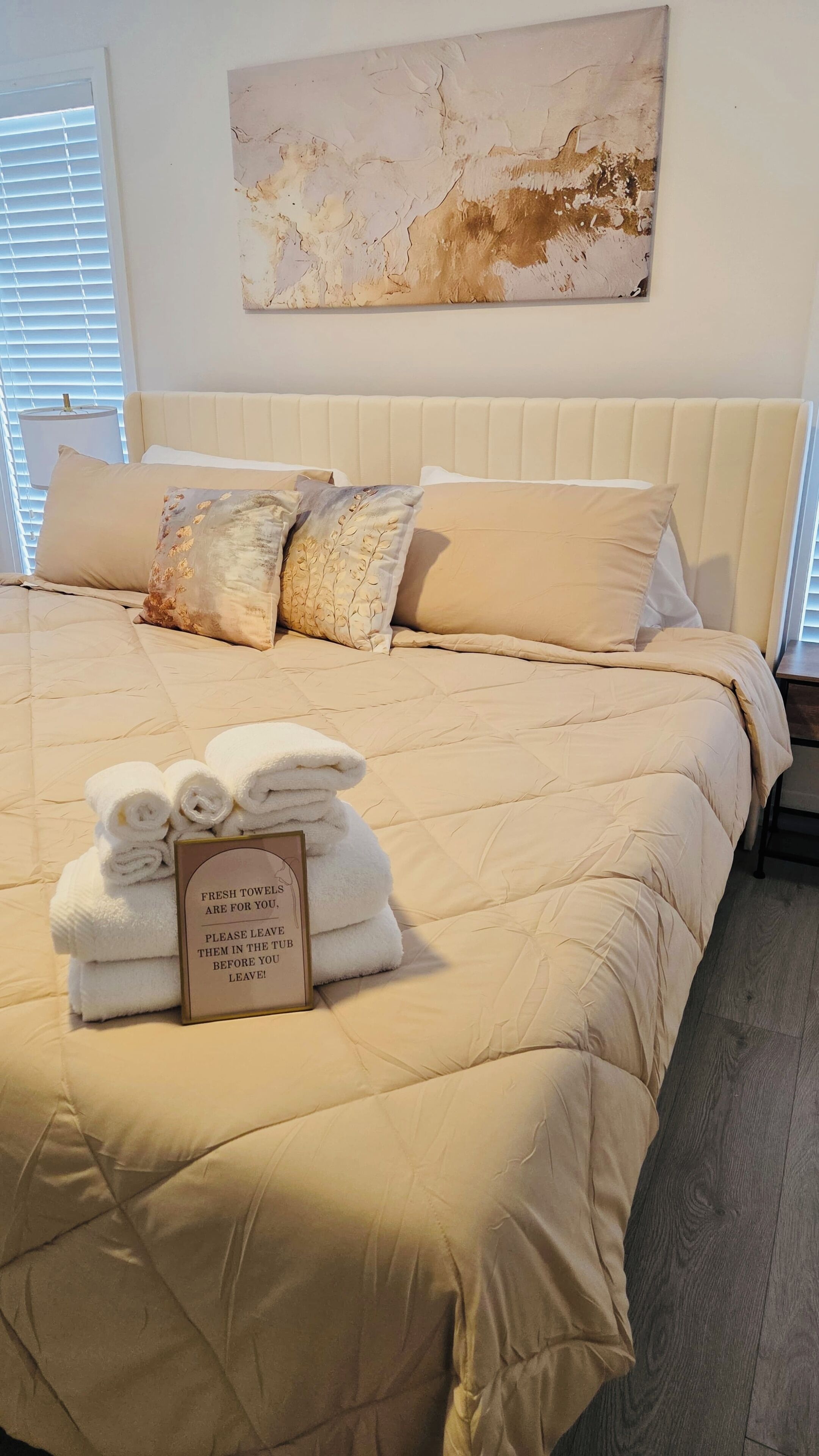 A neatly made bed with a beige comforter and pillows, a sign with fresh towels, and a large abstract painting on the wall.