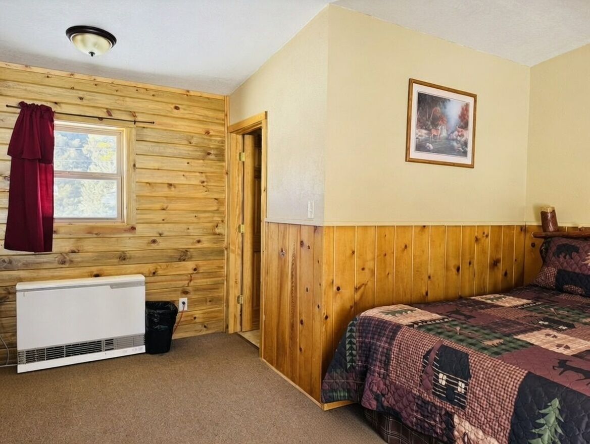A room with wooden paneling, a bed with a patterned quilt, a radiator, a window with curtains, and a framed picture on the wall.