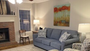 Uma sala de estar com um sofá azul, poltronas estampadas, uma pequena mesa de madeira, lareira e um ventilador de teto.