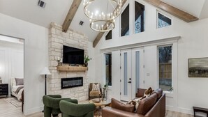 A spacious living room with a stone fireplace, a large TV mounted on the wall, and a high ceiling with exposed beams. - New! 4 BDRM Home near stockyards & DT Fort Worth (Fort Worth)