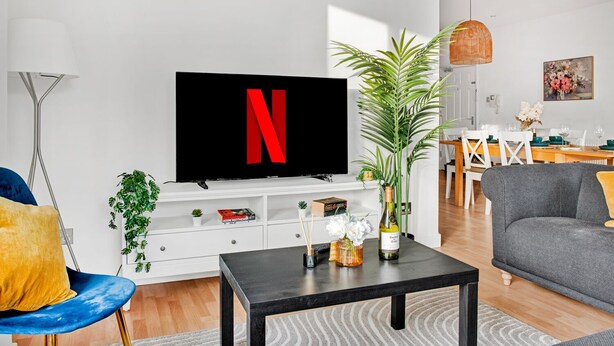 A modern living room with a flat-screen TV displaying a red 'N', a coffee table, a sofa, a dining area with a table and chairs, and a potted plant. - Spacious NG1 City Centre Apartment Near Motorpoint Arena, sleeps 6 (Nottingham)