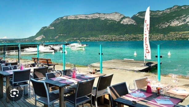 A seaside restaurant with outdoor seating overlooking turquoise waters and mountains. - Wonderful modern Chalet with private garden only 5 minutes walk to Annecy Lake. (Doussard)