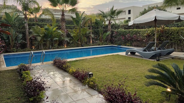 A swimming pool surrounded by lush greenery and palm trees, with lounge chairs and a white canopy. - Luxury 5-Bedroom Hotel Villa – Palm Valley Compound, Sheikh Zayed, Egypt (El Sheikh Zayed City)