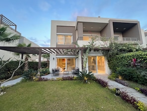 A modern two-story house with a patio area, outdoor seating, and lush landscaping.