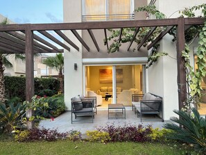 A covered outdoor seating area with two sofas and a table, surrounded by plants and a garden.