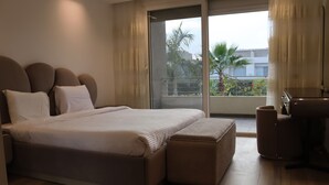 A modern bedroom with a large bed, a small seating area, and a balcony with a view of greenery. - Luxury 5-Bedroom Hotel Villa – Palm Valley Compound, Sheikh Zayed, Egypt (El Sheikh Zayed City)