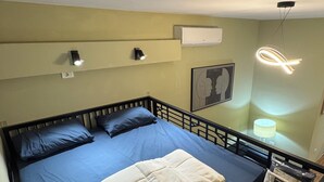 Ein Einzelbett mit blauer Matratze und weißen Kissen, ein Holzboden, ein schwarzes Metall-Kopfteil, eine Wand-Klimaanlage, eine Deckenleuchte und ein gerahmtes Kunstwerk an der Wand.