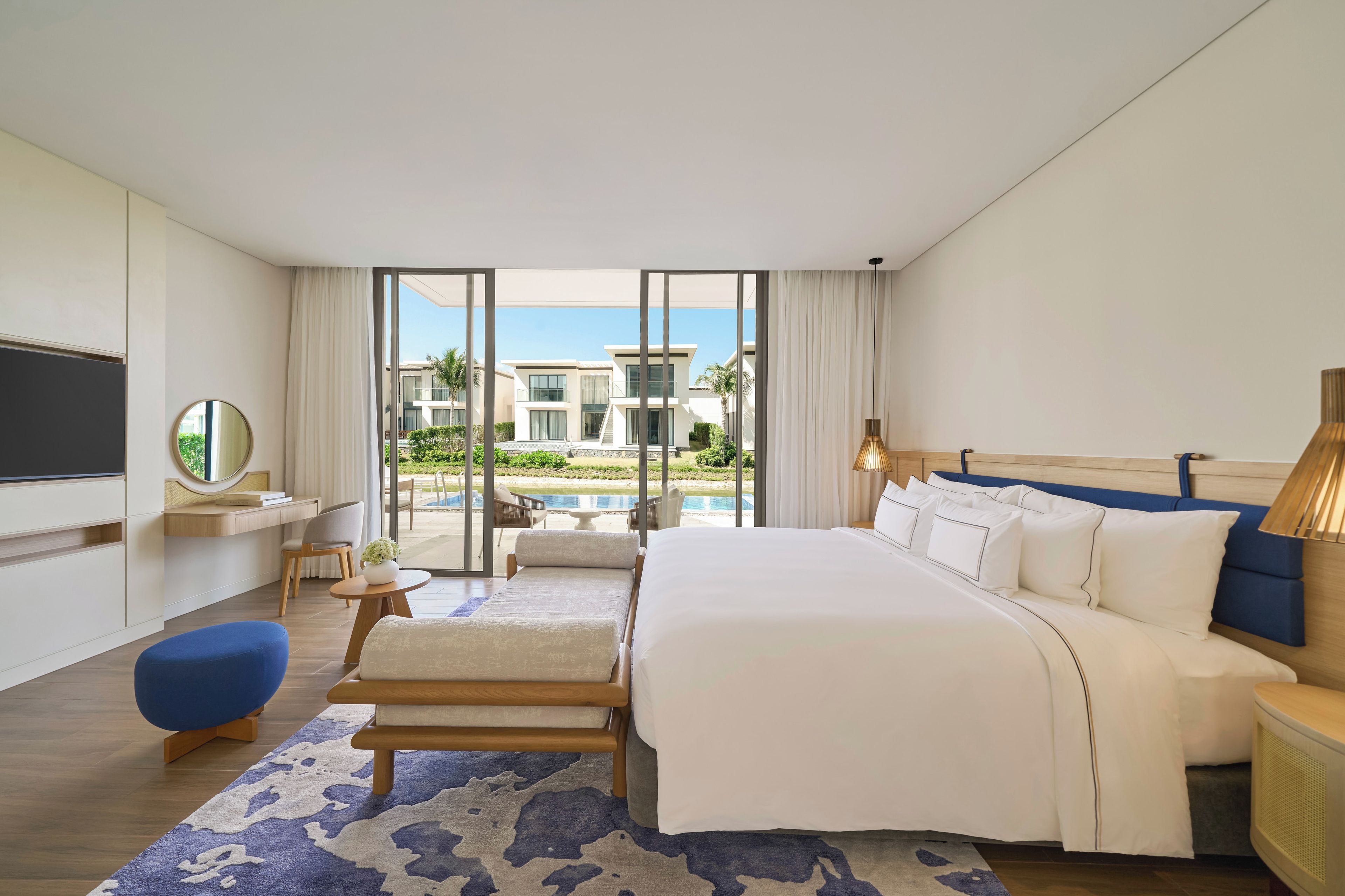 Habitación de hotel moderna con una cama grande, televisión, zona de estar y vistas a una piscina y edificios.