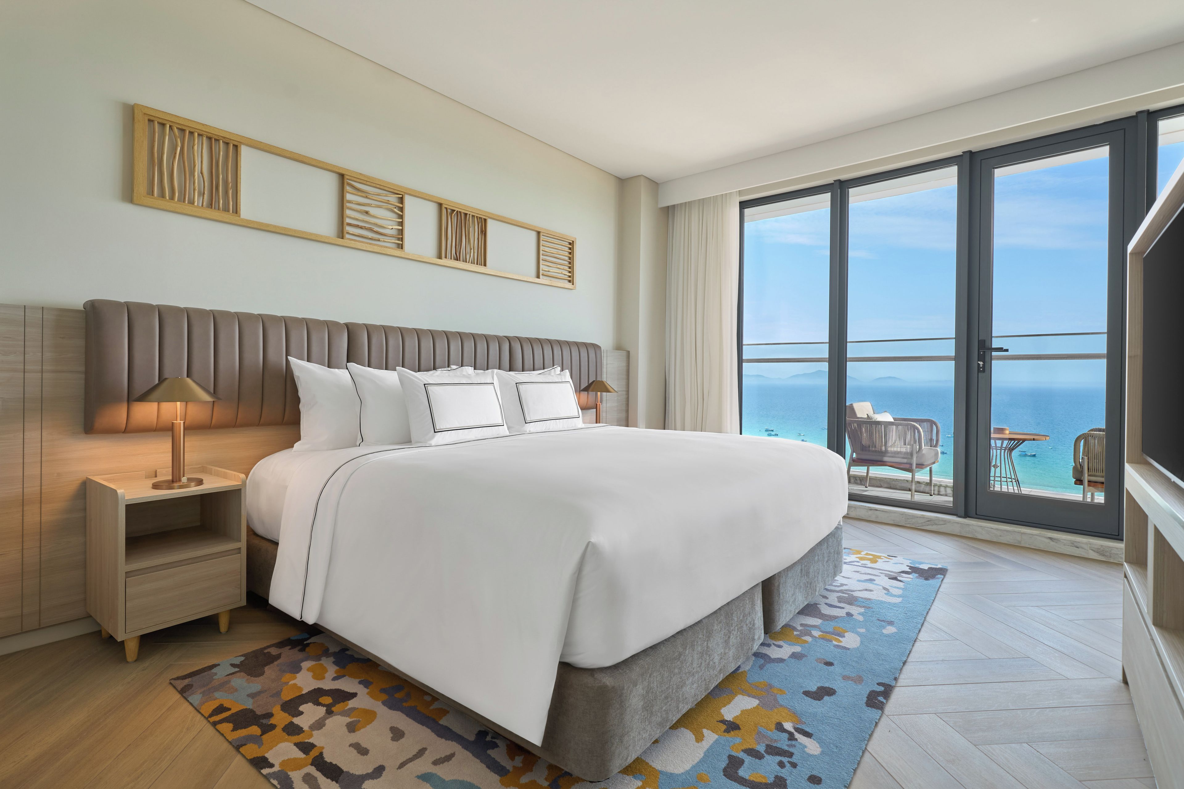 A modern hotel room with a large bed, a nightstand, a lamp, and a view of the sea through a balcony.
